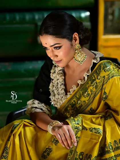 A downward-looking pose, highlighting the intricate patterns on the saree and the large, traditional earrings. The composition is elegant and focuses on the details of the styling.