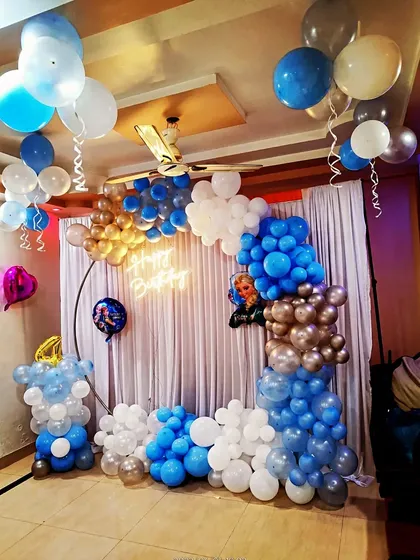 A simple and cozy Frozen-themed birthday setup at home. This decoration features a balloon arch in shades of blue and a neon 'Happy Birthday' sign against a white curtain backdrop.