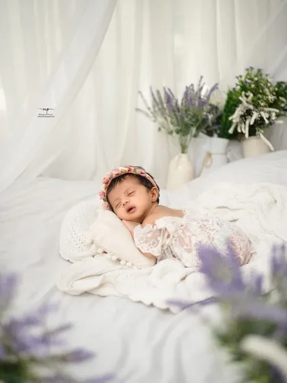 A newborn rests peacefully in a bed of soft white linens, framed by delicate lavender flowers. This lifestyle newborn photography approach creates a natural and timeless portrait.