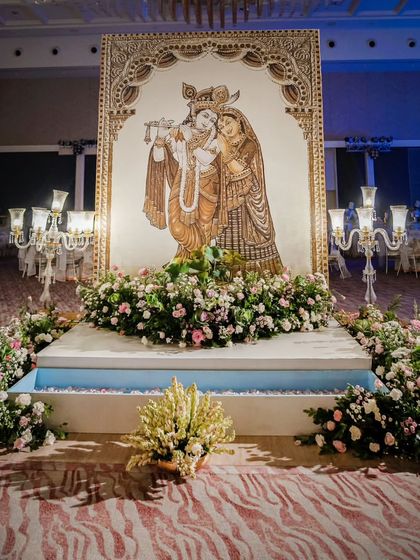 A beautiful photo backdrop featuring a large, hand-painted depiction of Radha and Krishna. The stage is set over a small water body and is adorned with lush floral arrangements and classic candelabras.