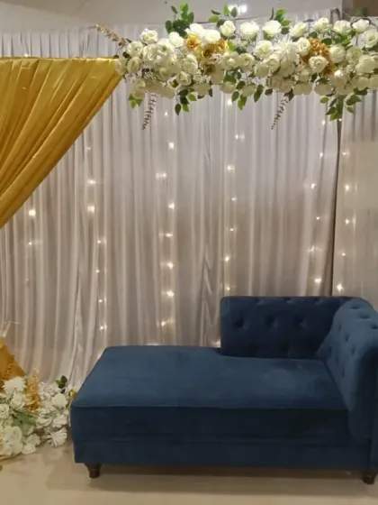 This engagement stage combines a white and gold floral arch with shimmering gold drapes and fairy lights. The setup includes floor candles and a royal blue sofa for a touch of color.