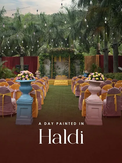 An overview of an outdoor Haldi ceremony setup. The aisle is lined with colorful seating leading to a beautiful mandap, all set against a backdrop of lush greenery and hanging lights for a magical feel.