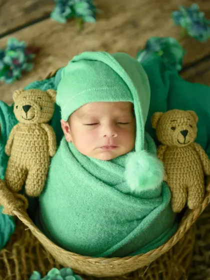 A full view of the teddy bear setup, showing the baby peacefully sleeping in a green pom-pom hat between two furry friends.
