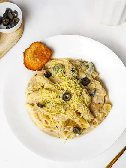 A bowl of creamy white sauce pasta topped with shredded cheese and black olives, with ingredients deconstructed on the side. This styled shot is great for a website or a feature menu item.