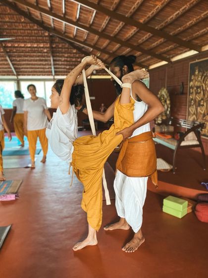 With the help of a strap, I assist a student in an advanced standing split variation. This demonstrates how we can work towards deeper flexibility with safety and support, honoring the body's current abilities while gently encouraging growth.