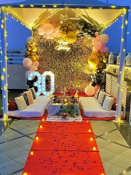 A glamorous 30th birthday party on a terrace, complete with a cabana, low seating for guests, and a red carpet entrance. The backdrop includes a gold shimmer wall, a 'Happy Birthday' neon sign, and marquee numbers.