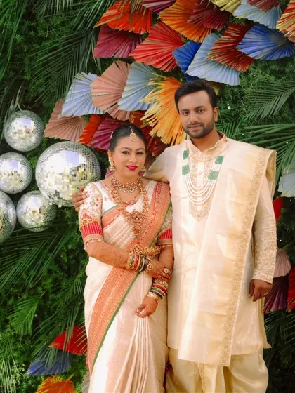 A full portrait of the wedding couple, showcasing their coordinated cream and red outfits against a festive backdrop. The details, from her bangles to his layered necklace, complete the picture-perfect look.