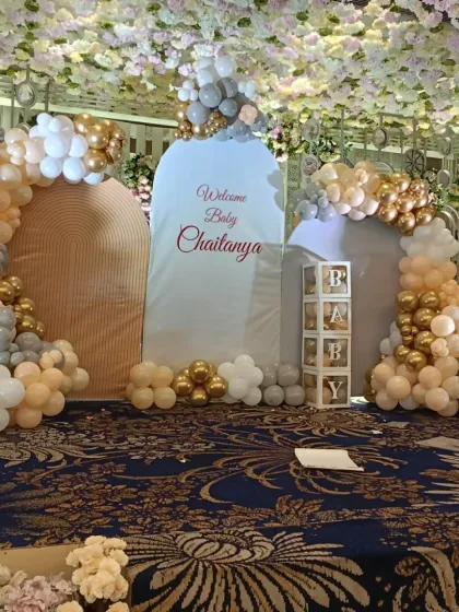 For grand celebrations in large halls, I design elaborate stage setups like this one for baby Chaitanya's welcome party. This features multiple backdrops, large organic balloon arches in gold, white, and beige, and transparent 'PARTY' boxes, all under a stunning floral ceiling.