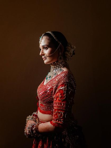 A classic, painterly portrait. The dramatic side-lighting sculpts the bride's features and showcases the rich texture and embroidery of her red lehenga.