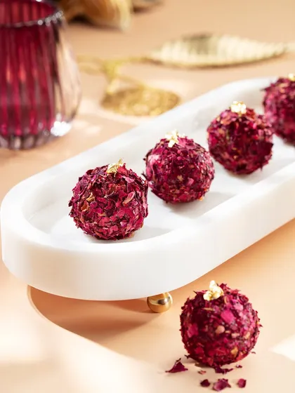 Rose-petal-covered laddoos from the Marriott Diwali collection, elegantly arranged on a white marble tray. The soft pink and gold tones in the background create a delicate and luxurious feel, perfect for festive gifting.