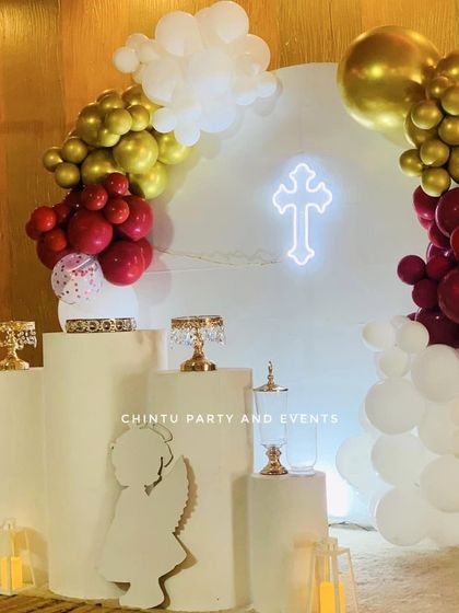A First Holy Communion decoration in a striking red, gold, and white color scheme. The setup features a circular backdrop with a neon cross, an angel cutout prop, and pedestals for cake and other items.