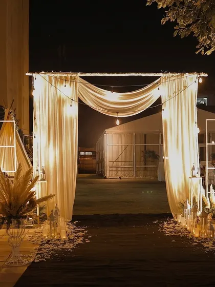 An ethereal entrance for a bohemian-themed night event. The path is lined with scattered petals and features a simple draped arch with warm Edison bulbs.