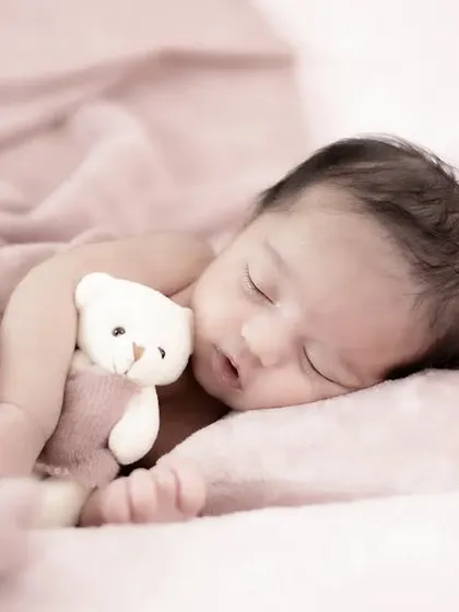A tender moment as a sleeping newborn cuddles a tiny teddy bear. The soft pink tones of the blanket and the gentle lighting create a warm and dreamy atmosphere.