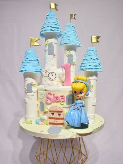 A fairytale first birthday cake designed as Cinderella's castle. This enchanting cake features blue turrets, a clock about to strike midnight, and a beautiful Cinderella figurine to complete the magical scene.