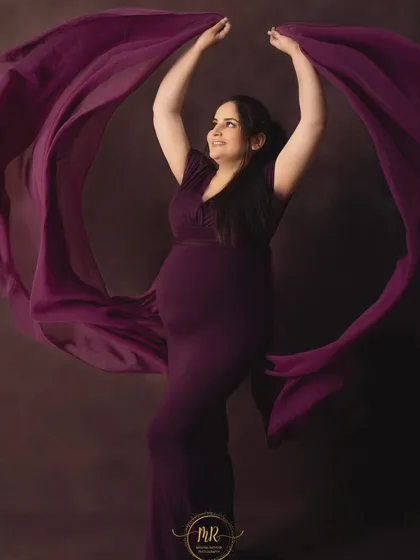 A joyful and dynamic solo portrait. The mother-to-be is wearing a deep purple gown, tossing the fabric in the air to create a beautiful, flowing shape around her.