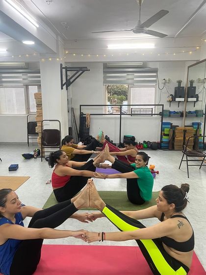 The studio buzzes with collaborative energy as students work together in partner boat pose variations.