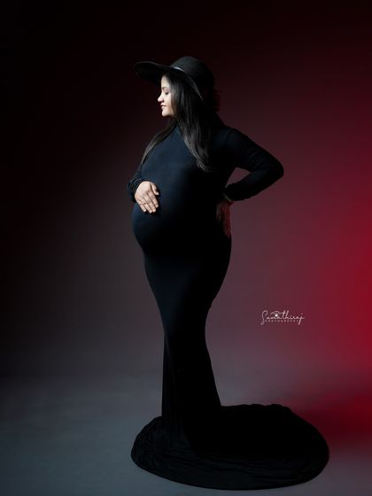 A dramatic portrait with colored backlighting. The mother-to-be, in a black gown and hat, strikes a powerful and stylish pose, creating a modern and artistic image.