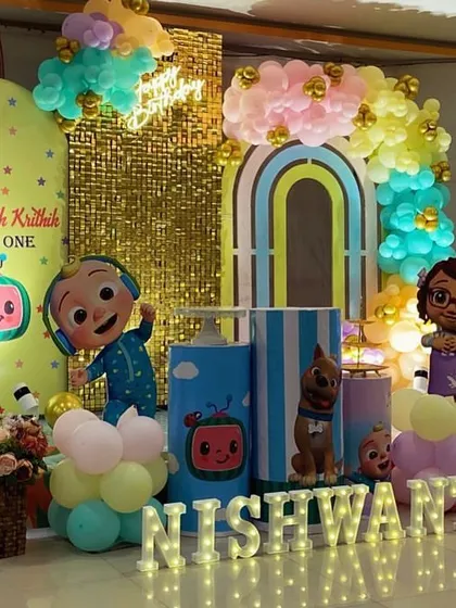 A bright and busy Cocomelon setup for Nishwanth, featuring a gold sequin wall, multiple character cutouts, and marquee letters.
