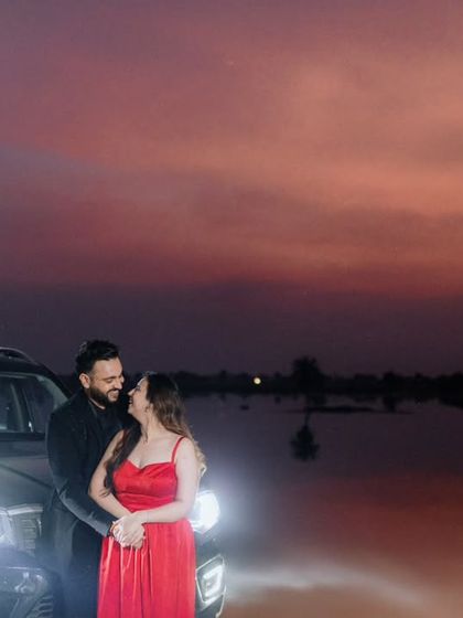A wide shot of a couple enjoying a romantic moment by a lake at dusk. The car's headlights illuminate them against the beautiful purple and orange sunset sky.