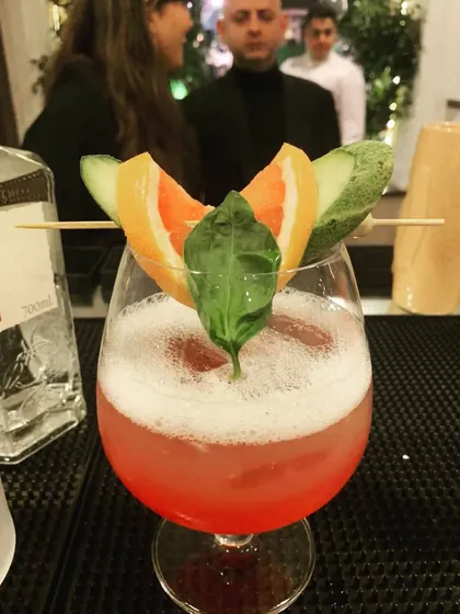 A beautifully garnished pink gin cocktail, ready to be served. The vibrant color, frothy top, and fresh garnishes of orange and basil showcase our attention to presentation and flavor.