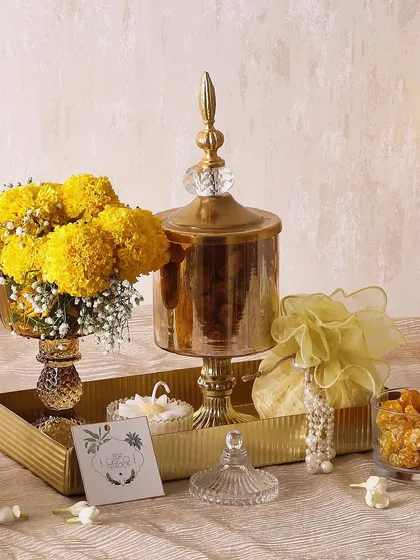 The Sweet Magnolia hamper is a beautiful arrangement of treats and decor. It includes a candle, trail mix, a flower vase with marigolds, and mini cookies, all set on a golden metal tray.