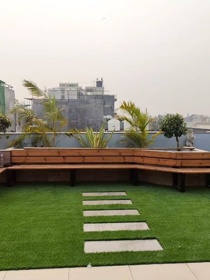 A custom-built L-shaped bench made from thermopine wood, set on an artificial lawn with a stone pathway. The bench is integrated with large planters.