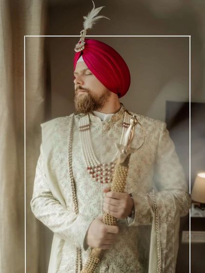 A regal portrait of the groom, Samuel, in his ivory sherwani and pink turban, holding his kirpan, looking out a window.