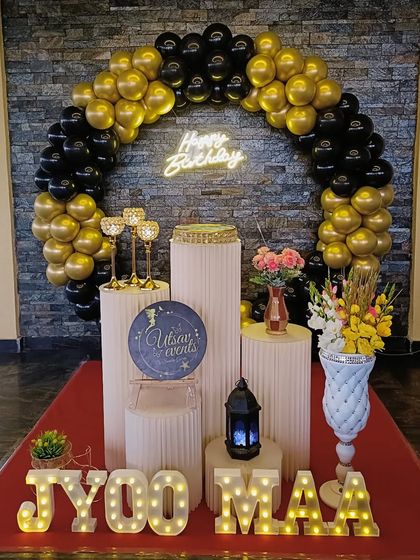 A stylish black and gold balloon arch against a stone wall backdrop. This setup includes marquee letters and elegant plinths for a modern birthday celebration.