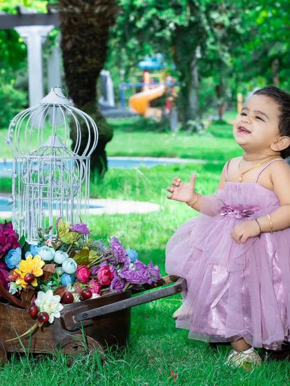 A beautiful outdoor shot of a baby girl in a purple dress, looking up with a joyful expression next to a cart of flowers.