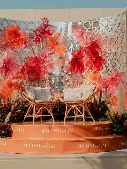 The complete stage view, featuring rattan chairs, a mirrored jaali backdrop, and an explosion of colorful paper flowers.