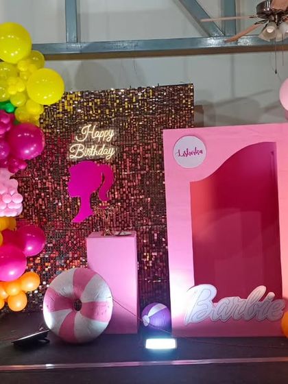 A vibrant, tropical Barbie-themed birthday party. This setup features a pink "doll box" photo booth, a sequin backdrop, and colorful balloon clusters with palm tree cutouts.