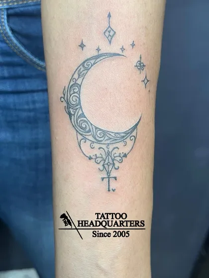 An ornamental crescent moon tattoo on the forearm, featuring intricate swirls and sparkling star accents for a magical look.