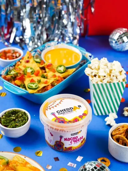 A party-ready spread featuring nachos with Cheddar Together cheese dip, popcorn, and other snacks. The vibrant blue background and confetti create a fun and festive atmosphere for New Year's.