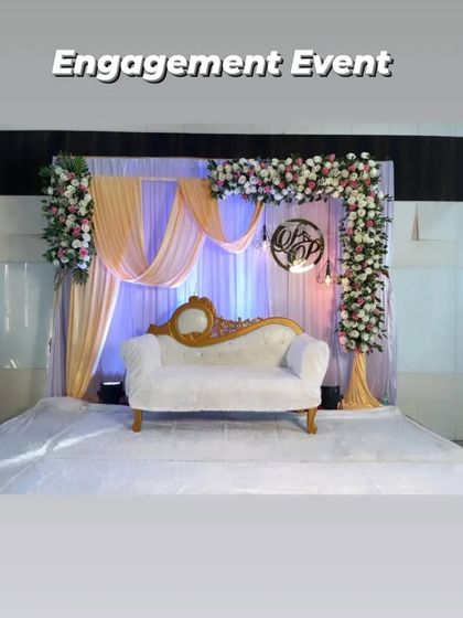 A complete view of an elegant engagement stage. The setup includes a floral arch, soft peach and white draping, a custom monogram, and a classic white sofa, all professionally lit to create a warm and inviting atmosphere.