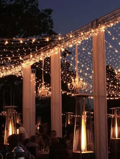An outdoor evening event illuminated by a canopy of fairy lights. The setup includes white pillars, crystal chandeliers, and patio heaters, creating a warm and magical ambiance for a dinner or reception.