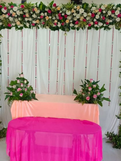 A simple and elegant stage decoration suitable for a small function or ceremony. The setup features a fresh floral arch made of white and pink roses over a clean white curtain backdrop.