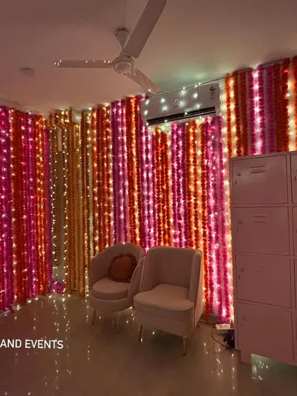 A photo-ready seating area created for a home Mehendi party. The wall is covered in vertical garlands of pink, red, and orange marigolds, interwoven with fairy lights for a magical glow.