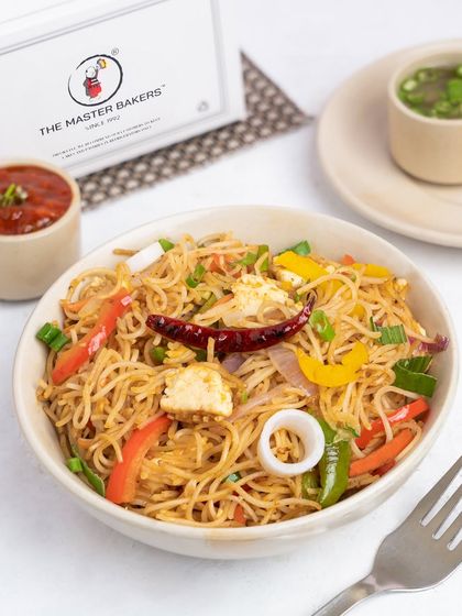A bowl of our Chili Garlic Noodles, tossed with colorful bell peppers, onions, and chunks of paneer. The dish is garnished with a whole dried red chili for an extra kick.