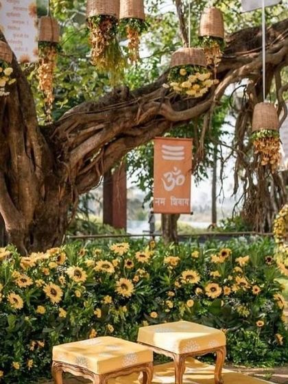 A rustic Haldi setup under a large banyan tree, with hanging floral baskets and a sunflower-covered seating area.