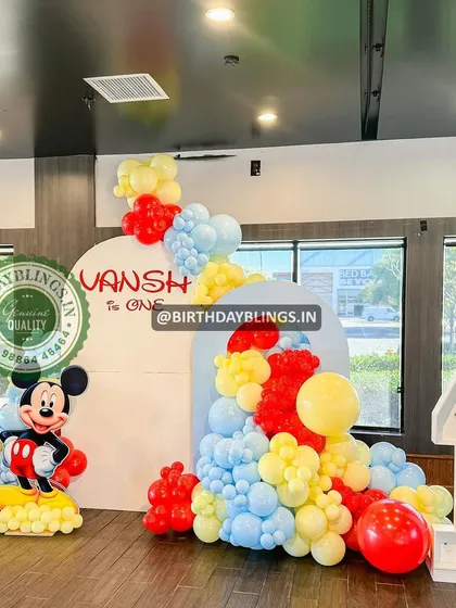 A bright and cheerful Mickey Mouse first birthday party. The setup includes a classic Mickey cutout, a light-up number '1', and a balloon cascade in red, yellow, and blue.