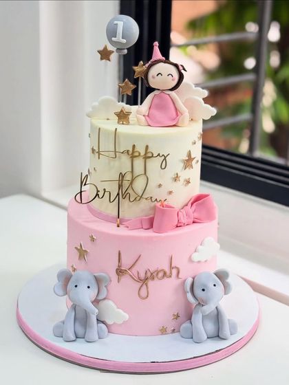 Turning ONE never looked so dreamy! This two-tier cake for a first birthday features adorable fondant elephants, a princess topper, and a soft pink and white color palette.