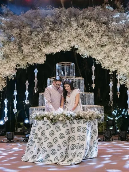 A picture perfect moment from a grand sangeet. The couple cuts their cake under a massive floral arch, framed by hanging crystals and dramatic cold pyro effects for that ultimate wow-factor.