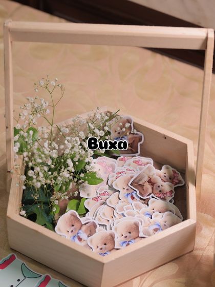 Preparing the elements for a teddy bear baby shower, including custom stickers and fresh flowers in a hexagonal pinewood basket.