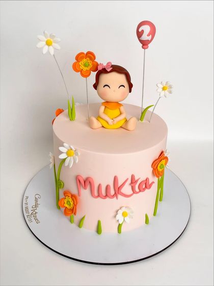 A full view of the floral-themed cake for Mukta's second birthday, showcasing the simple pink frosting adorned with a cheerful baby topper and whimsical flowers.