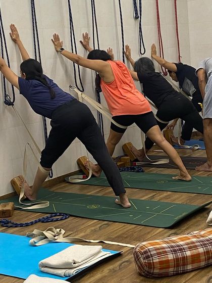 A group of students practicing Parsvottanasana (Pyramid Pose) at the wall. They are using blocks and belts to maintain alignment and deepen the forward bend.
