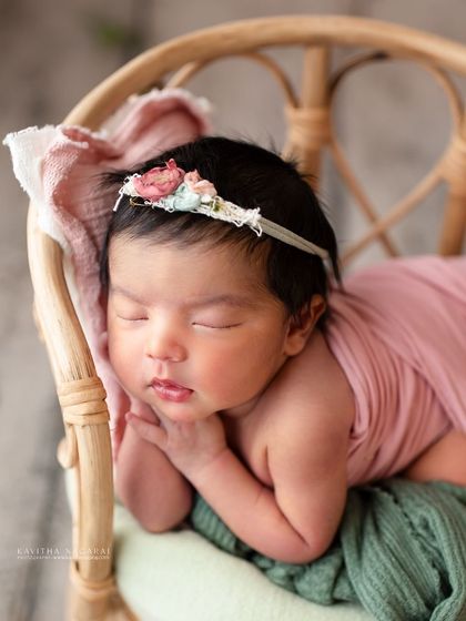 A nine-day-old newborn girl resting in a miniature wicker chair. The soft pink and green tones and floral headband create a delicate, dreamy portrait.