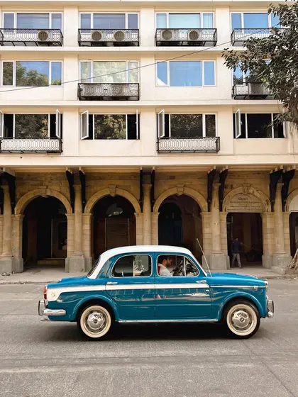A blue and white Fiat, another classic, driving past the arched windows of a heritage building. This series celebrates the timeless appeal of vintage cars in Mumbai.