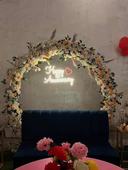 A beautiful setup for an anniversary celebration. The decor features a floral arch, a 'Happy Anniversary' neon sign, and a comfortable blue sofa.