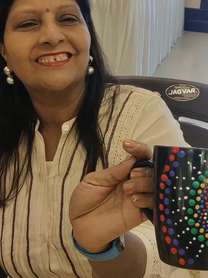 A smiling woman from the Sakhi Ladies Group holds up a black ceramic mug she has beautifully decorated with a colourful dot mandala pattern. This showcases a popular activity for adult group sessions and kitty parties, combining socializing with a relaxing craft.