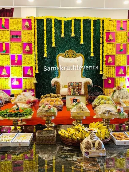 A vibrant Seemantha backdrop in yellow and pink with hanging marigold strings. The foreground shows a lavish display of covered offering trays and gift boxes, ready for the ceremony.